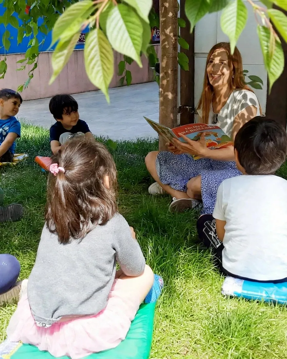 Ağaç gölgesinde çimlerde oturan çocuklara kitap okuyan öğretmen